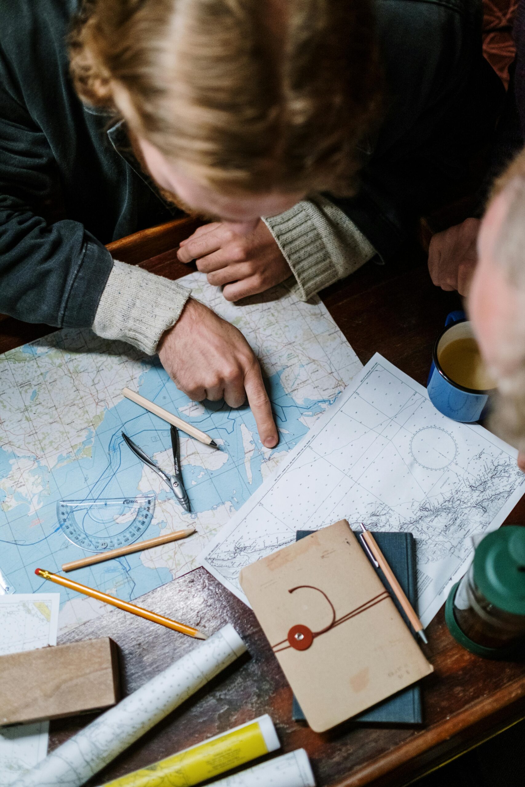 Two adults planning a journey using nautical maps and tools on a wooden table.